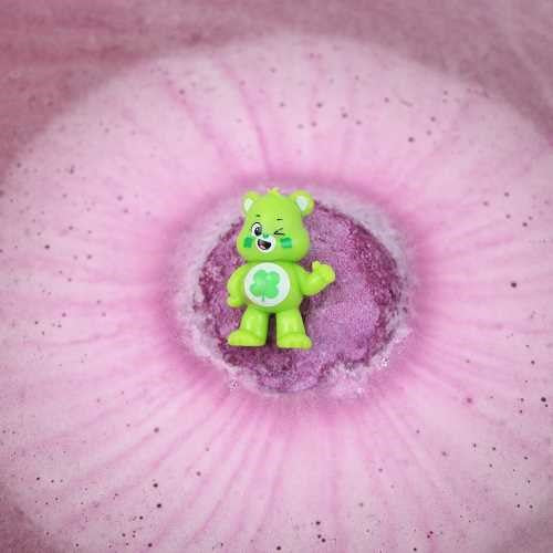 Figura verde do Ursinho Carinhoso com trevo de quatro folhas ao centro de uma bomba de banho roxa efervescente.