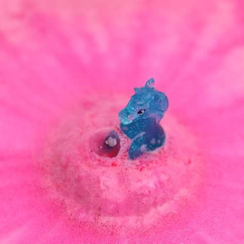 Figura de unicórnio azul brilhante a emergir de uma bomba de banho rosa efervescente na água.