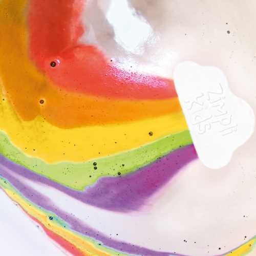 Detalhe da bomba de banho Zimpli Kids arco-íris a dissolver na água, libertando cores vibrantes.