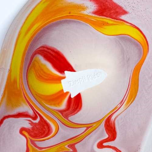 Vista aproximada da ZIMPLIKIDS Bomba de banho Foguetão a efervescer na água, criando um turbilhão de cores vibrantes de vermelho e amarelo.