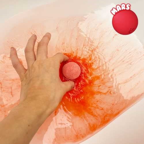 Mão a colocar a bomba de banho Marte ZIMPLIKIDS na água, criando uma efervescência avermelhada.
