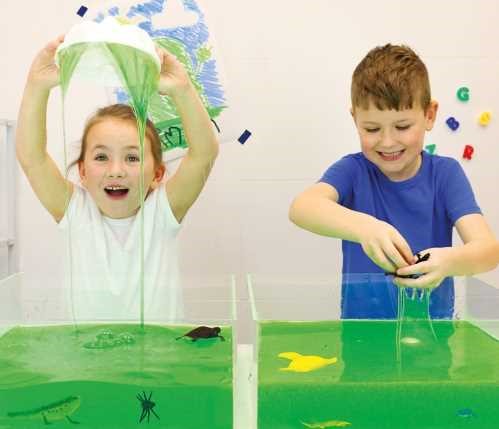 Duas crianças a interagir com o ZIMPLIKIDS Slime para banho Baff Verde em recipientes transparentes. Uma criança segura o slime a pingar, enquanto a outra explora o material com brinquedos.