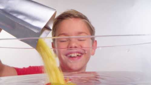 Criança sorridente a deitar pó amarelo de uma saqueta Zimplikids Crackle Baff Colours em água, criando uma experiência divertida e colorida.