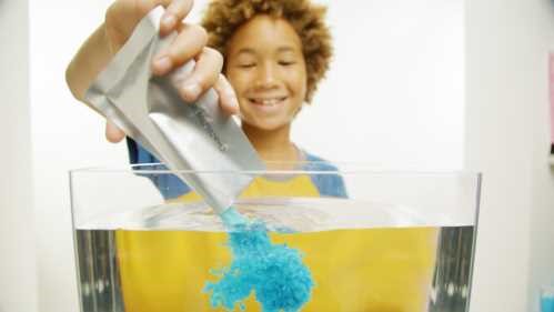 Criança sorridente a deitar pó azul de uma saqueta Zimplikids Crackle Baff Colours em água, com o pó a espalhar-se na água.