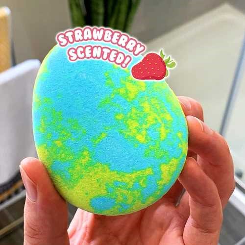 Detalhe da bomba de banho ZIMPLIKIDS Tartaruga 100gr na mão, mostrando a sua forma oval e as cores azul e verde, com um autocolante "Strawberry Scented!".