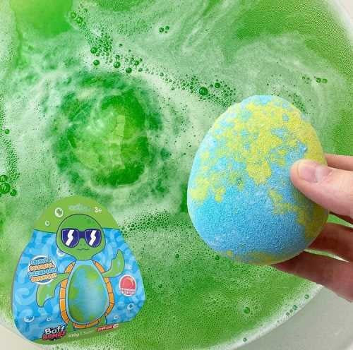 Bomba de banho ZIMPLIKIDS Tartaruga 100gr prestes a ser colocada numa banheira com água verde já efervescente, mostrando a embalagem ao lado.