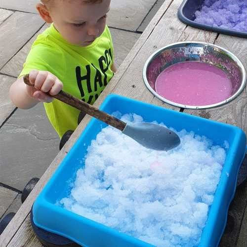 Criança pequena a mexer neve artificial ZIMPLIKIDS colorida numa bandeja, explorando a textura e o jogo sensorial.