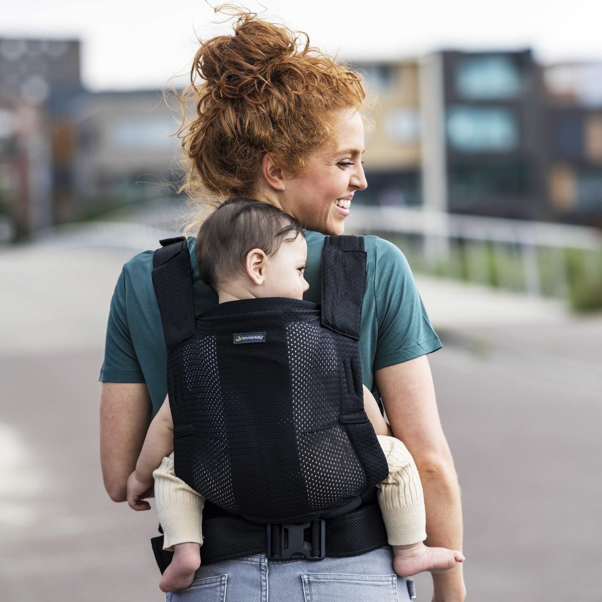 Mulher a utilizar a Mochila Porta-bebés Minimonkey com um bebé pequeno nas costas, em ambiente urbano.