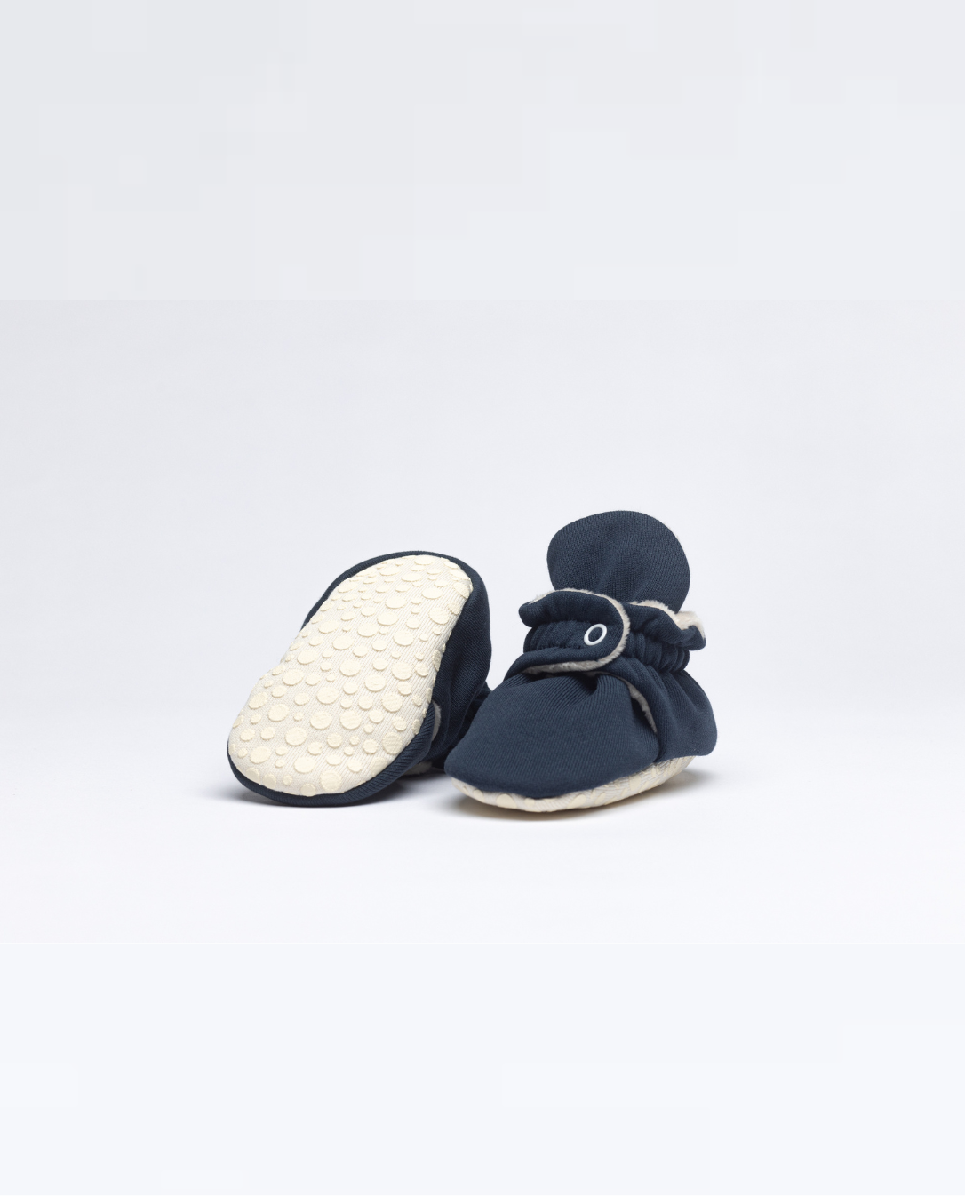 Pantufas pretas com solas expostas, destacando o seu design antiderrapante e adaptável para bebés que começam a caminhar.
