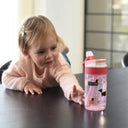 Uma criança pequena, com cabelo loiro e camisola rosa, a alcançar a Garrafa Kambukka LAGOON Pink Flamingo 400ml numa mesa escura.