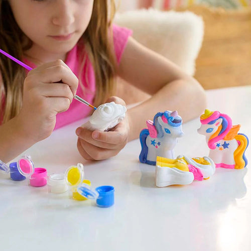 Criança a pintar um dos unicórnios de gesso ainda branco com um pincel fino, enquanto as outras figuras terminadas estão na mesa.
