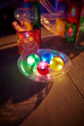 Quatro bolas luminosas (verde, azul, rosa e amarela) dentro de uma pequena taça transparente, posicionadas numa pista de bolas desfocada ao fundo.