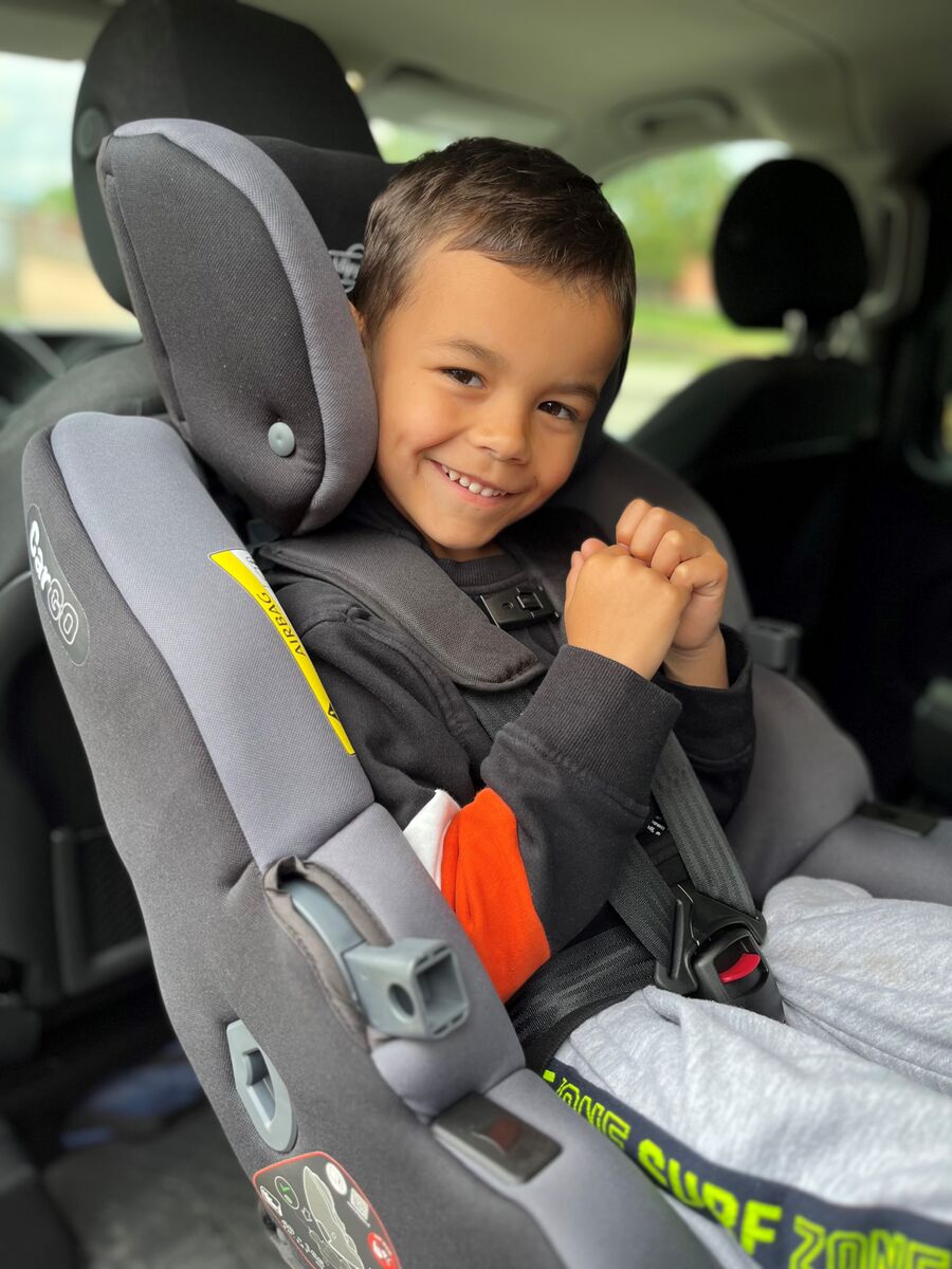 Criança sorridente sentada na cadeira auto Klippan CarGO instalada contra marcha no carro, destacando o conforto e a segurança do arnês de 5 pontos.