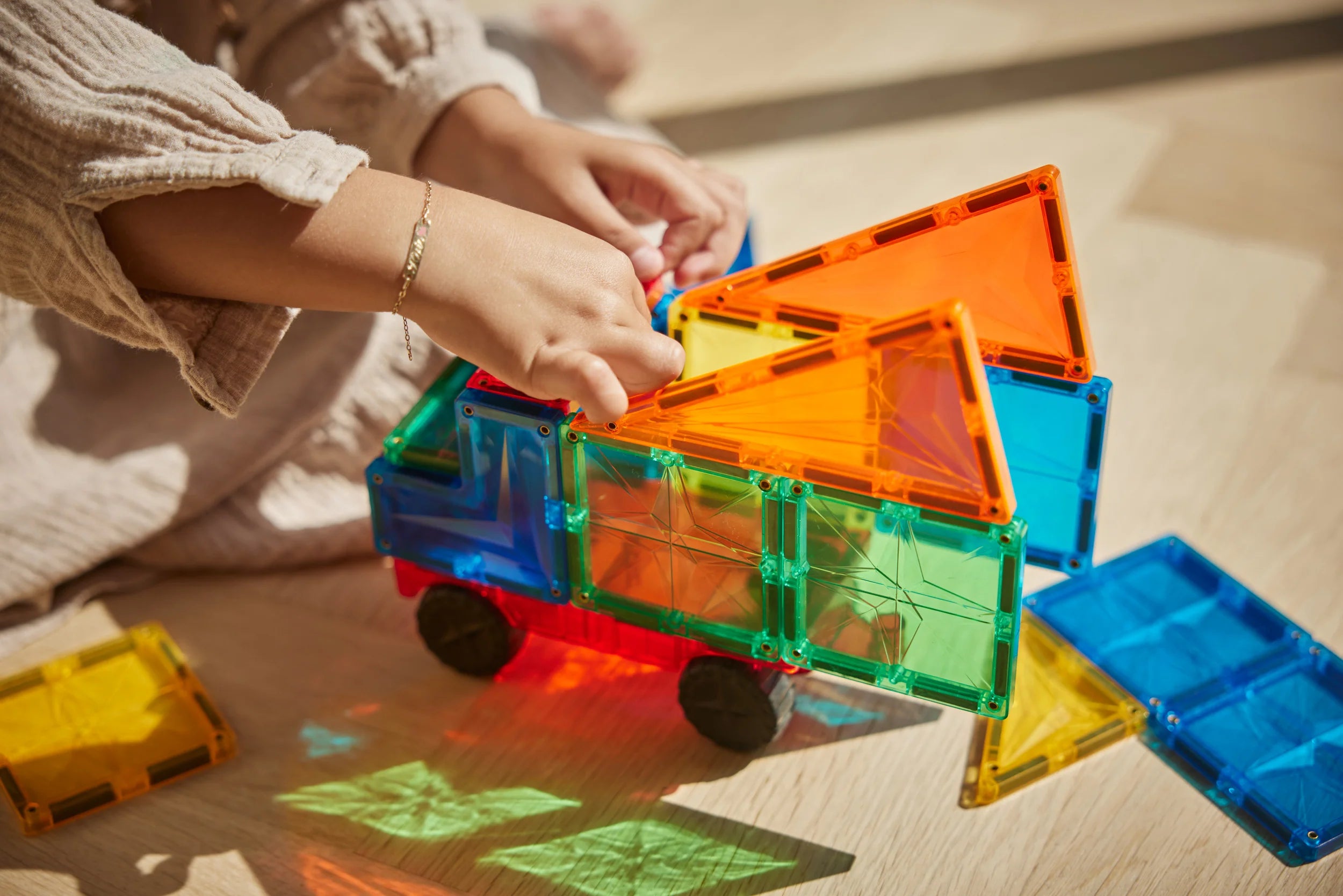 Mãos de uma criança a encaixar peças magnéticas coloridas e transparentes para construir a cabina de um camião de brincar, com luz solar a incidir no chão.