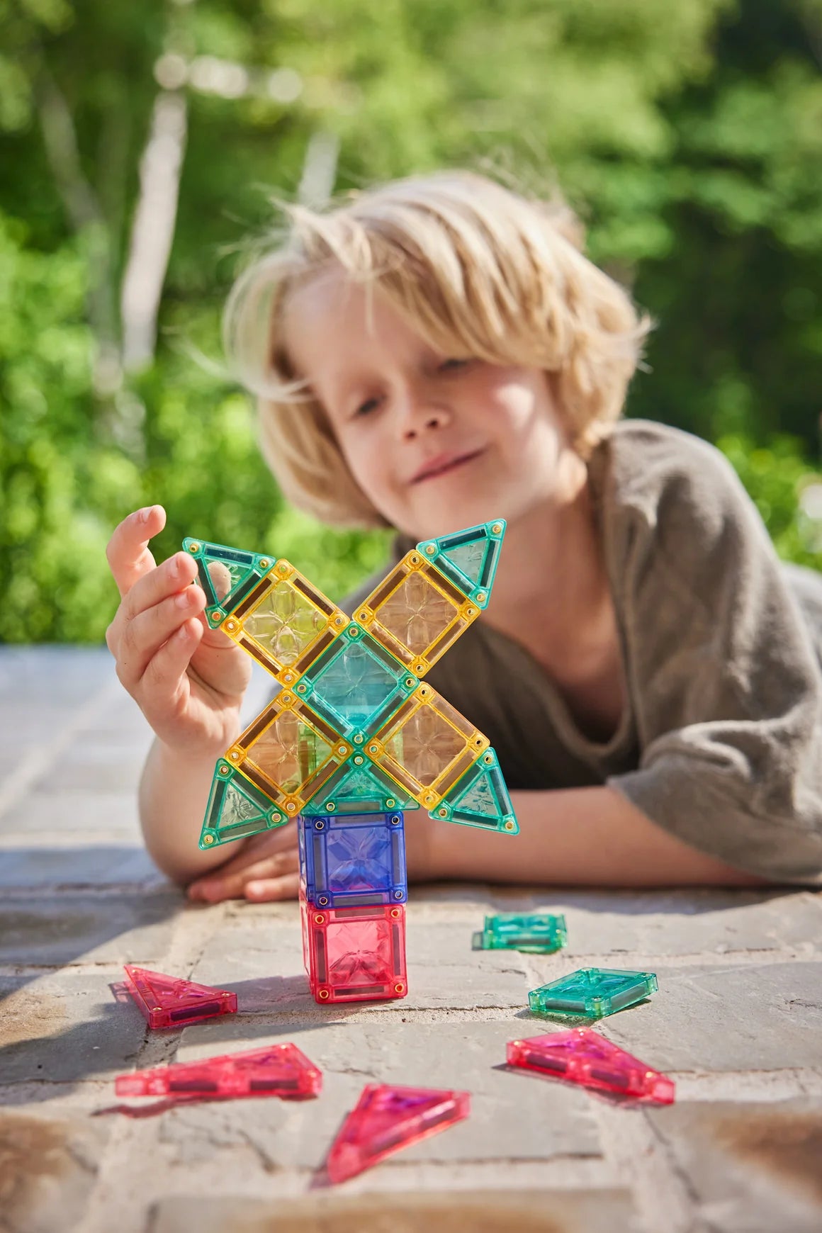 Uma criança loira a brincar com blocos magnéticos Clever Clixx em tons pastel ao ar livre, construindo uma estrutura.