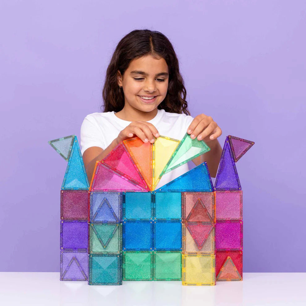 Menina sorridente a montar um castelo colorido e brilhante com os blocos magnéticos Connetix Glitter Castle Pack.