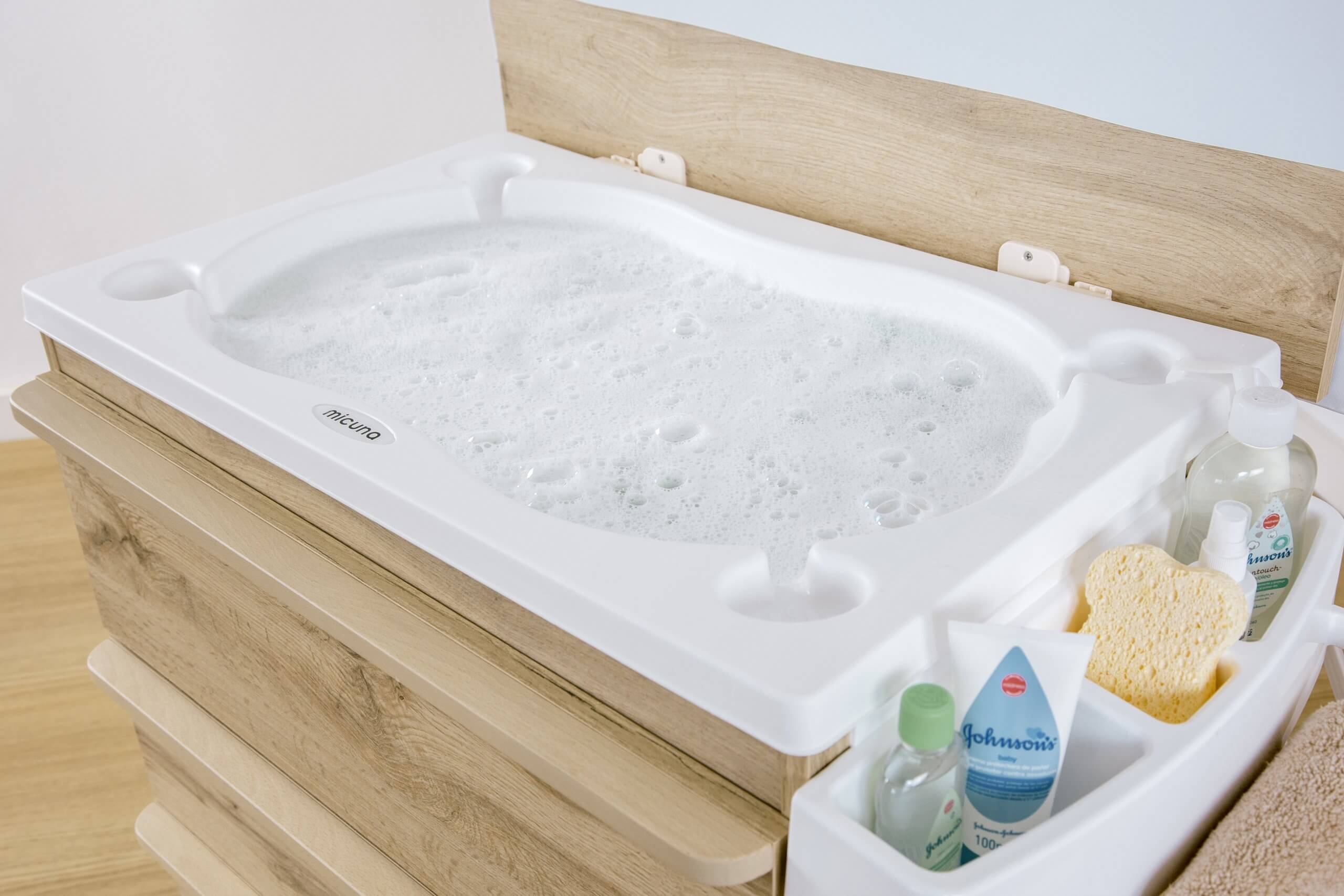 Detalhe da cuba de banho da Micuna Martha Móvel Banheira cheia de espuma, com produtos de higiene de bebé no organizador lateral.
