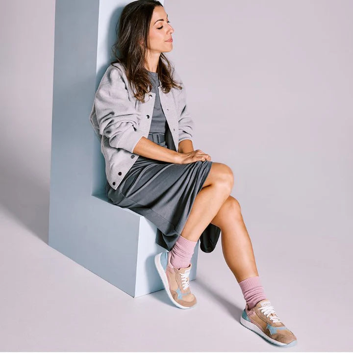 Mulher com cabelo castanho e roupa cinza sentada, exibindo as sapatilhas MUM Barefoot Daniela Cloud nos pés, com meias rosa.