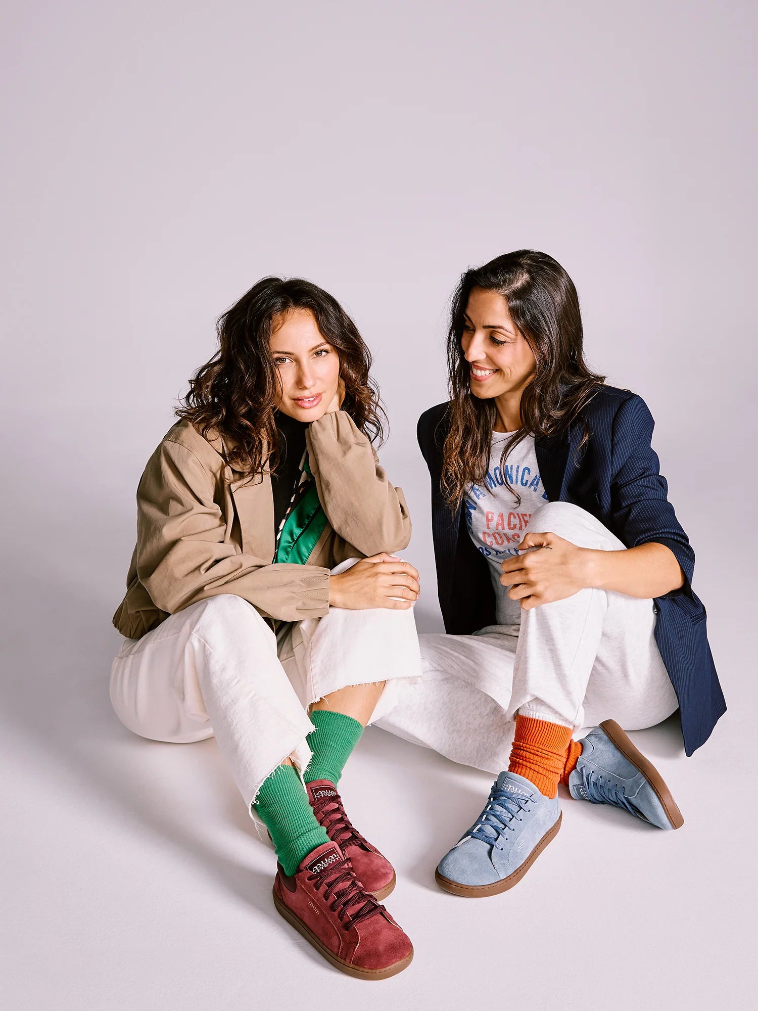 Duas mulheres sentadas, uma a usar sapatilhas MUM Barefoot Diana Wine bordô e outra sapatilhas azuis.