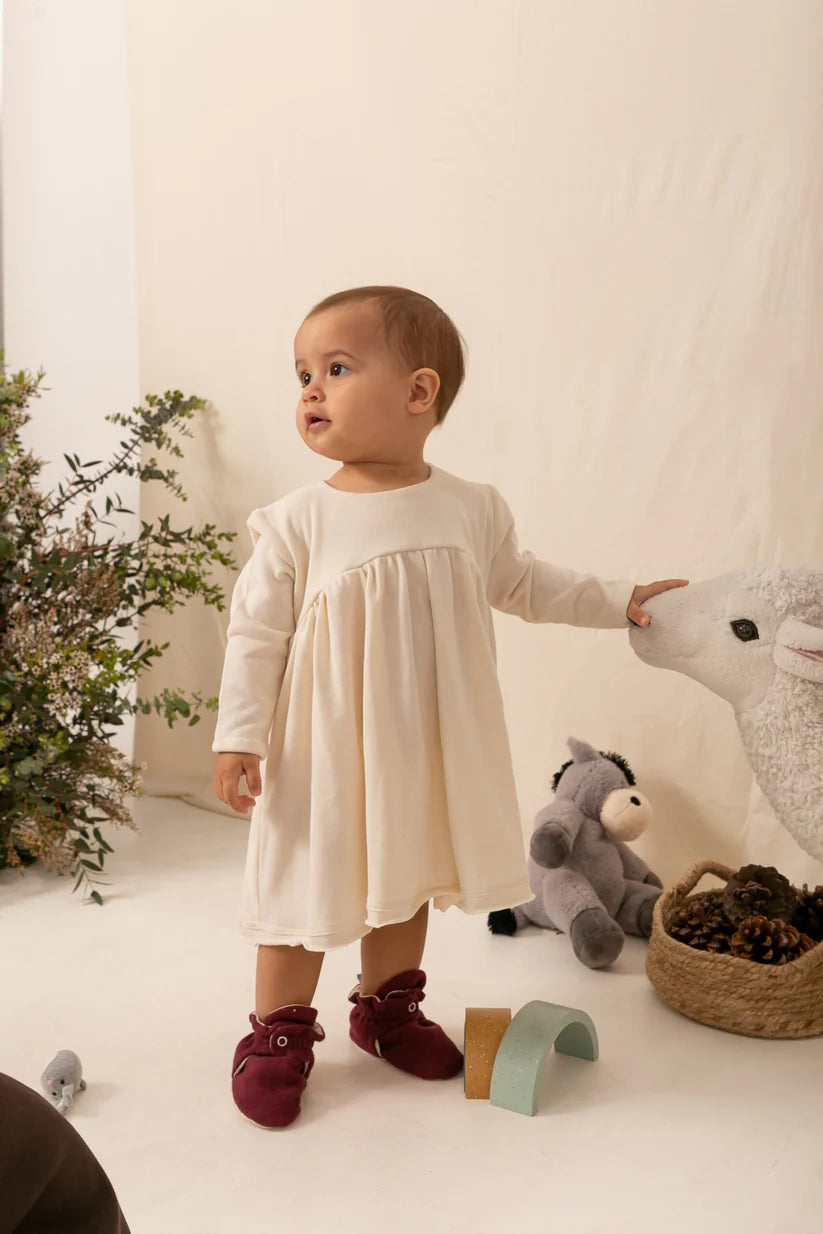 Bebé com vestido creme e pantufas botinhas Zás Trás Cherry Pie, em pé, olhando para a direita, num ambiente acolhedor com brinquedos e plantas.