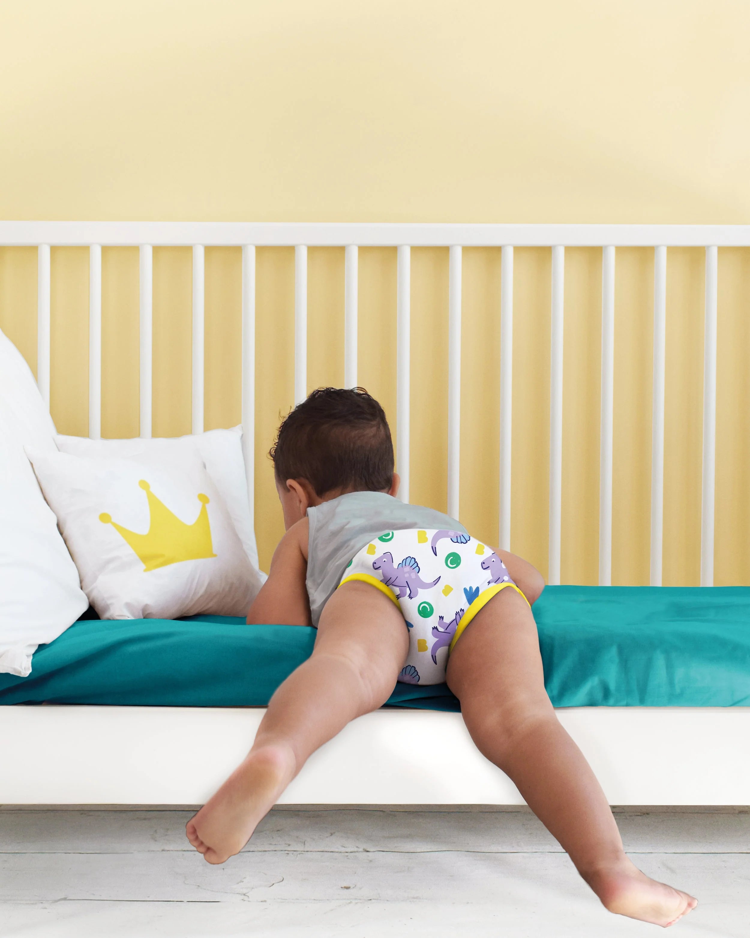 Criança de costas com cueca de treino Bambino Mio de dinossauros, deitada na cama, a explorar o ambiente.