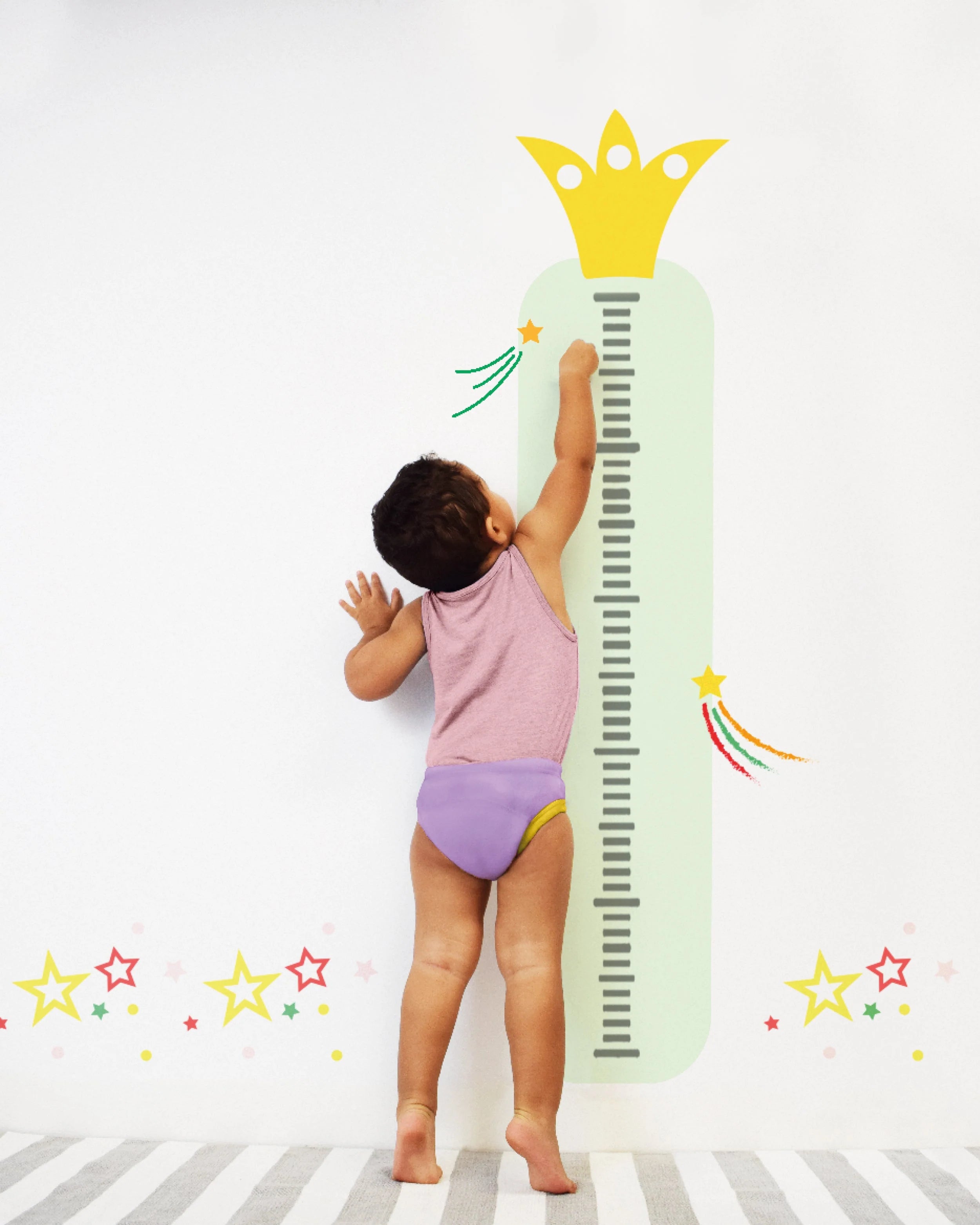 Criança em cueca de treino Bambino Mio roxa, descalça, a medir a altura numa parede decorada.