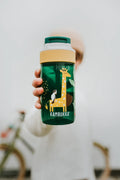 Garrafa Kambukka LAGOON Safari Jungle 400ml, verde escura com design de girafa e tucano, sendo segurada por uma criança desfocada.