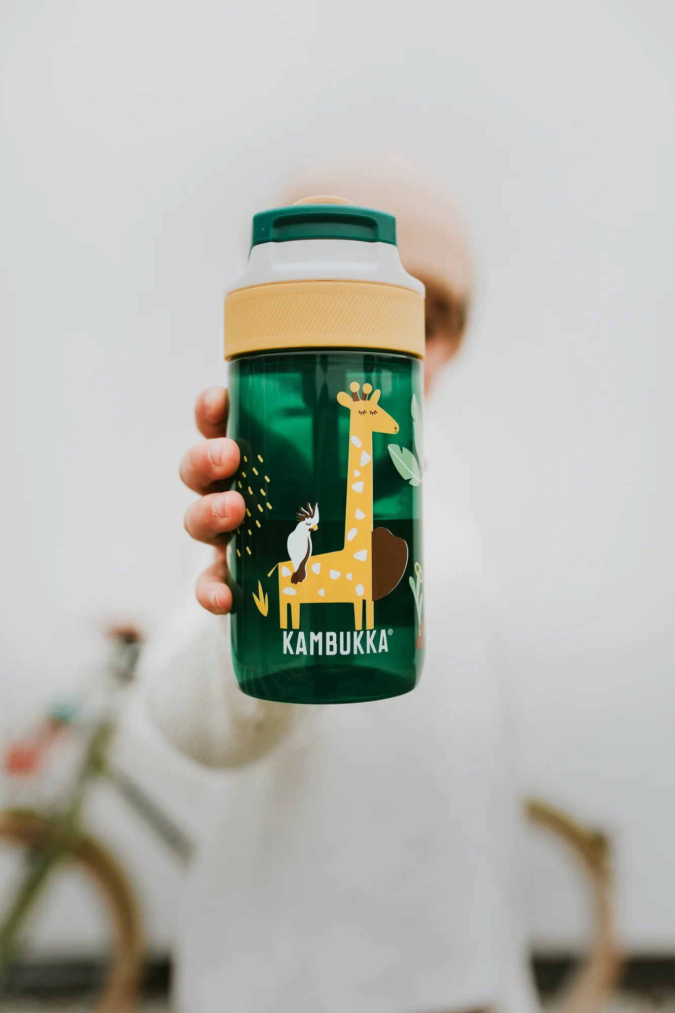 Garrafa Kambukka LAGOON Safari Jungle 400ml, verde escura com design de girafa e tucano, sendo segurada por uma criança desfocada.