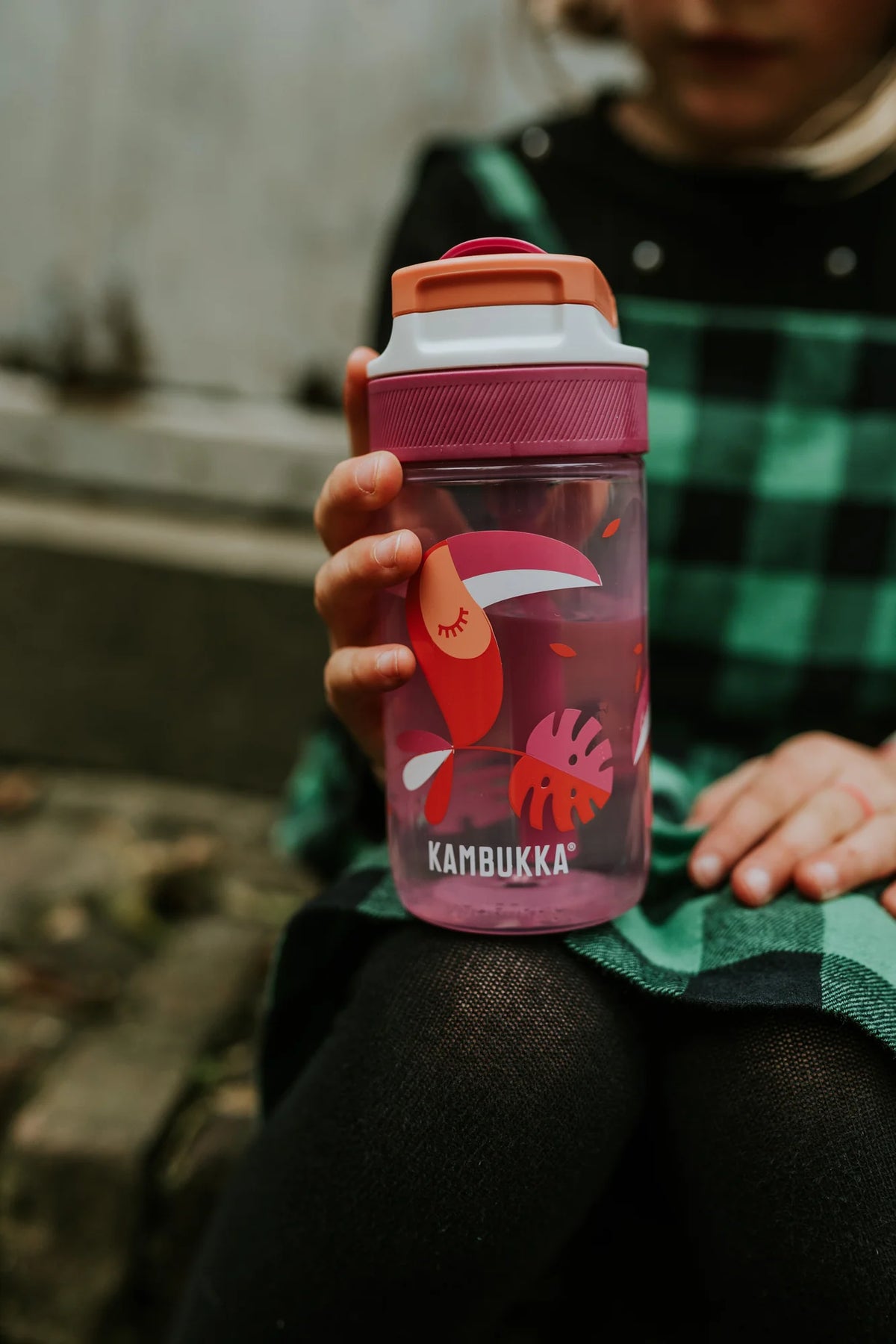 Criança sentada ao ar livre segurando a garrafa Kambukka LAGOON Toekan Love 400ml rosa com design de tucano, cheia de água.