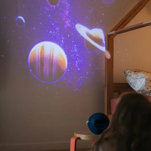 Projeção de planetas e galáxias no teto e parede de um quarto infantil, com o projetor KidyStar visível na mesa de cabeceira.