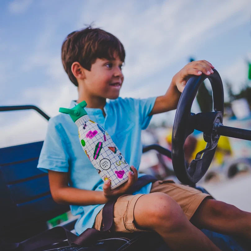 Criança sorridente a segurar a Garrafa Térmica Cool Bottles Play Mode 500ml enquanto brinca com um volante, em ambiente exterior.
