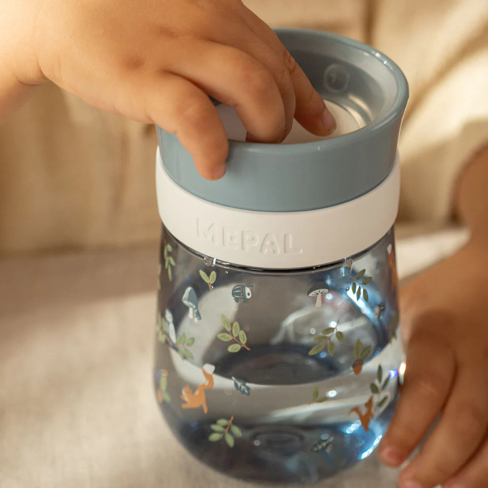 Mãos de criança a segurar o copo de treino Little Dutch Mepal 360º Forest Friends, com a tampa aberta e líquido visível.
