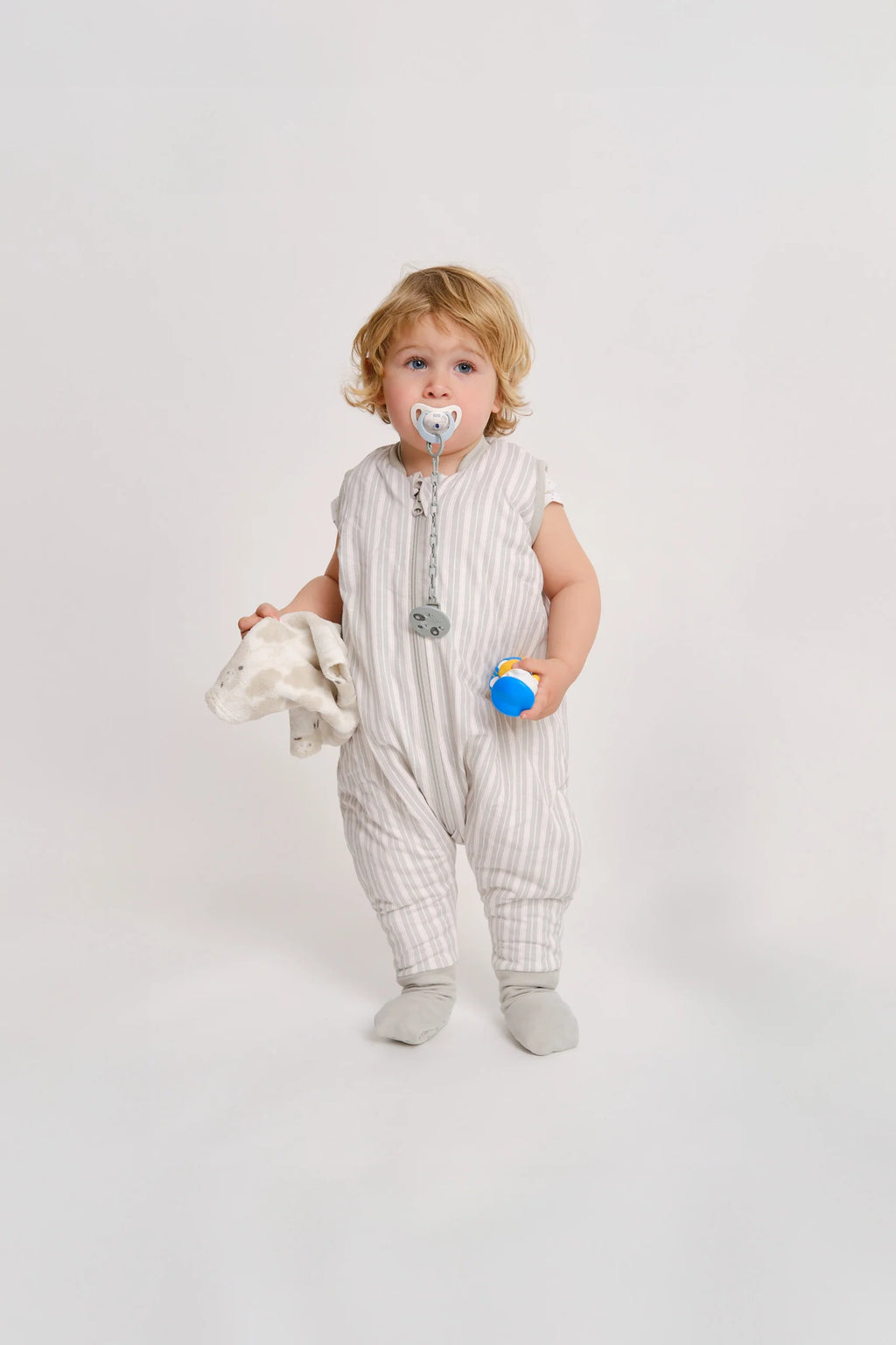 Bebé loiro com chupeta, de pé, a usar o saco de dormir MolisandCo TOG 1.0 Dawn Stripes com pés e meias, segurando um pano e um brinquedo.