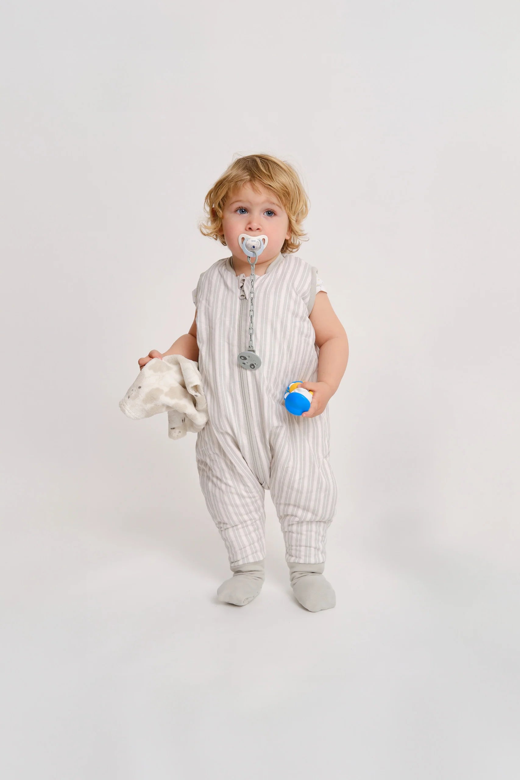 Bebé loiro com chupeta, de pé, a usar o saco de dormir MolisandCo TOG 1.0 Dawn Stripes com pés e meias, segurando um pano e um brinquedo.