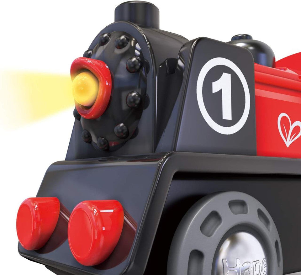 Detalhe da frente do Hape Comboio com Luz a Pilhas, destacando a luz amarela acesa e os pormenores do design preto e vermelho.