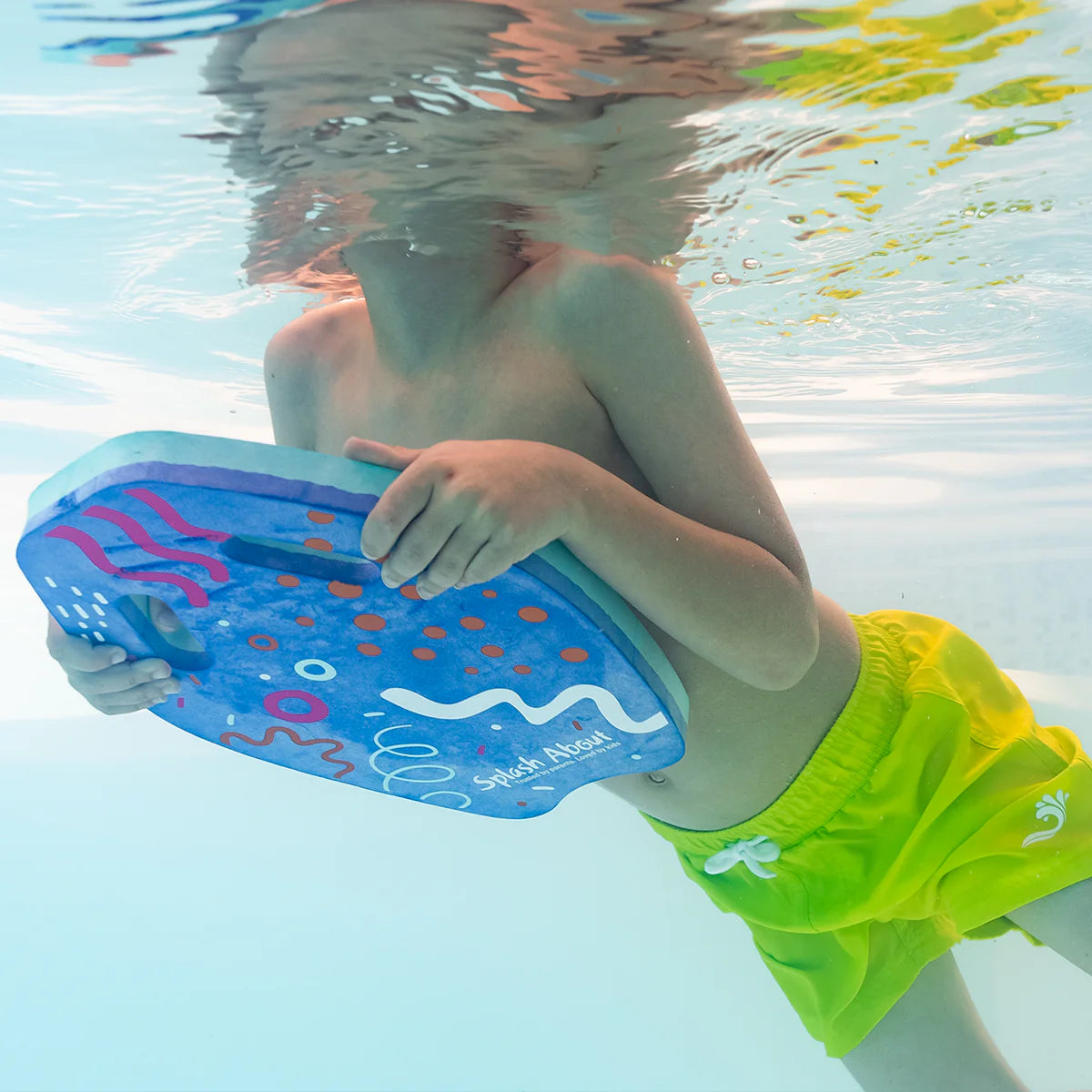 Criança submersa na piscina, a segurar com as duas mãos uma prancha de natação Splash About - Kickboard Junior azul com padrões coloridos, focando-se na aprendizagem.