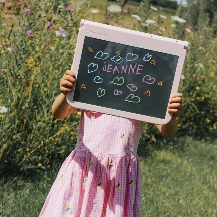 Criança ao ar livre, vestida com um vestido às bolinhas rosa, a segurar a prancha de desenho KidyDraw rosa onde escreveu "Jeanne" com corações desenhados.