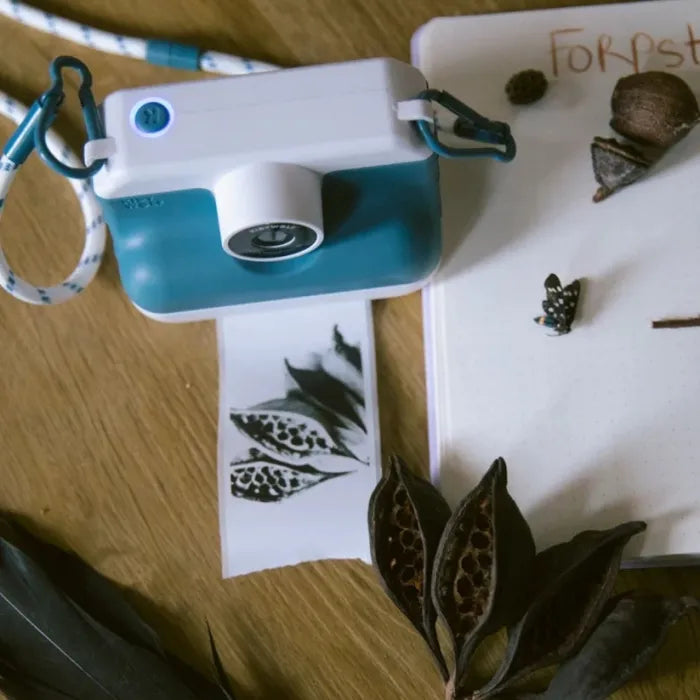 Máquina fotográfica KidyWolf K-PRINT azul e branca sobre uma superfície de madeira, ao lado de um caderno com a palavra 'Forest' e elementos naturais secos.