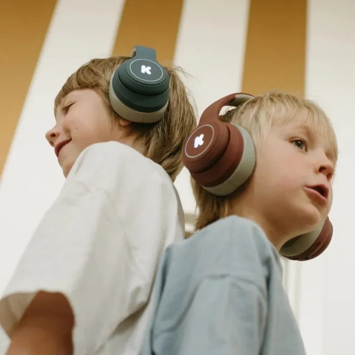 Duas crianças sorridentes, um rapaz e uma rapariga, a usar auscultadores KidyWolf K-Ears em tons de verde-azeitona/castanho, com fundo de riscas amarelas e brancas.