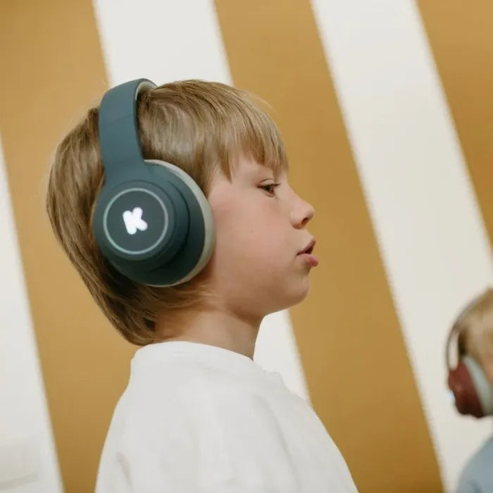 Close-up do perfil de um rapaz a usar os auscultadores KidyWolf K-Ears verde-azeitona, com fundo de riscas amarelas e brancas, focado no uso.