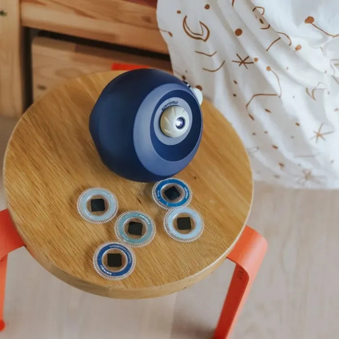 Vista superior do projetor KidyStar azul pousado num pequeno banco de madeira, com os seis discos de projeção ao lado.