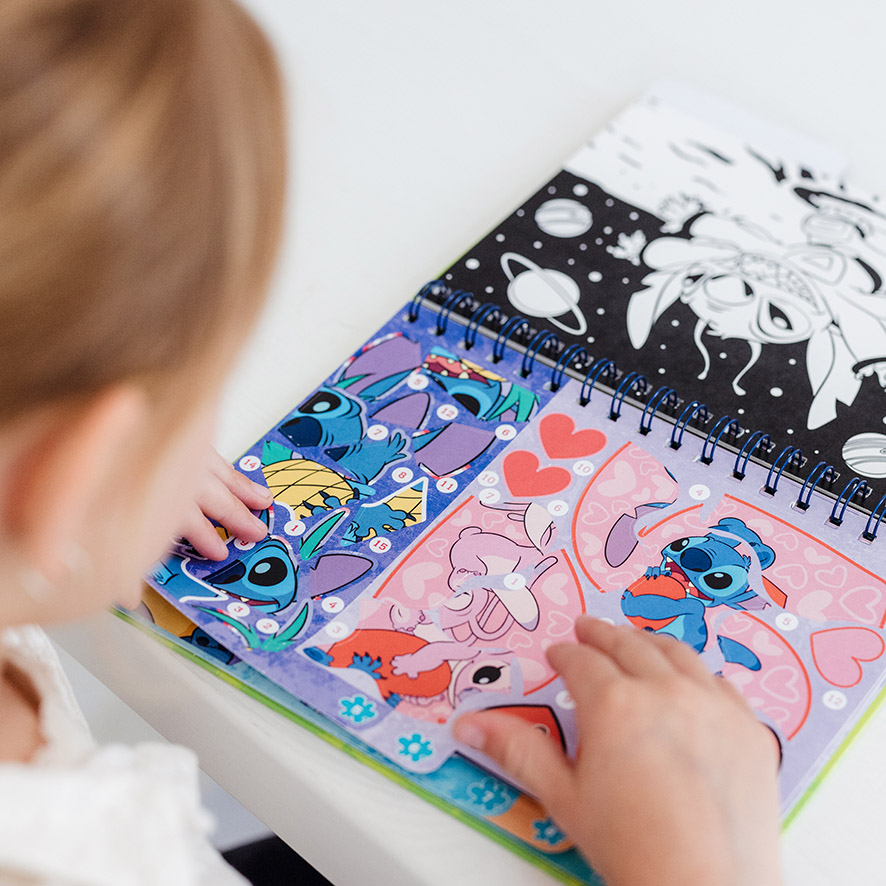 Mãos de criança a interagir com o livro Totum Sticker by Number Stitch, colocando autocolantes nas áreas numeradas.