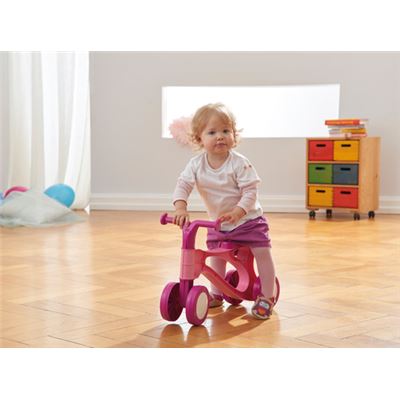 Uma menina pequena a andar na scooter de equilíbrio cor-de-rosa dentro de casa, sobre pavimento de madeira.