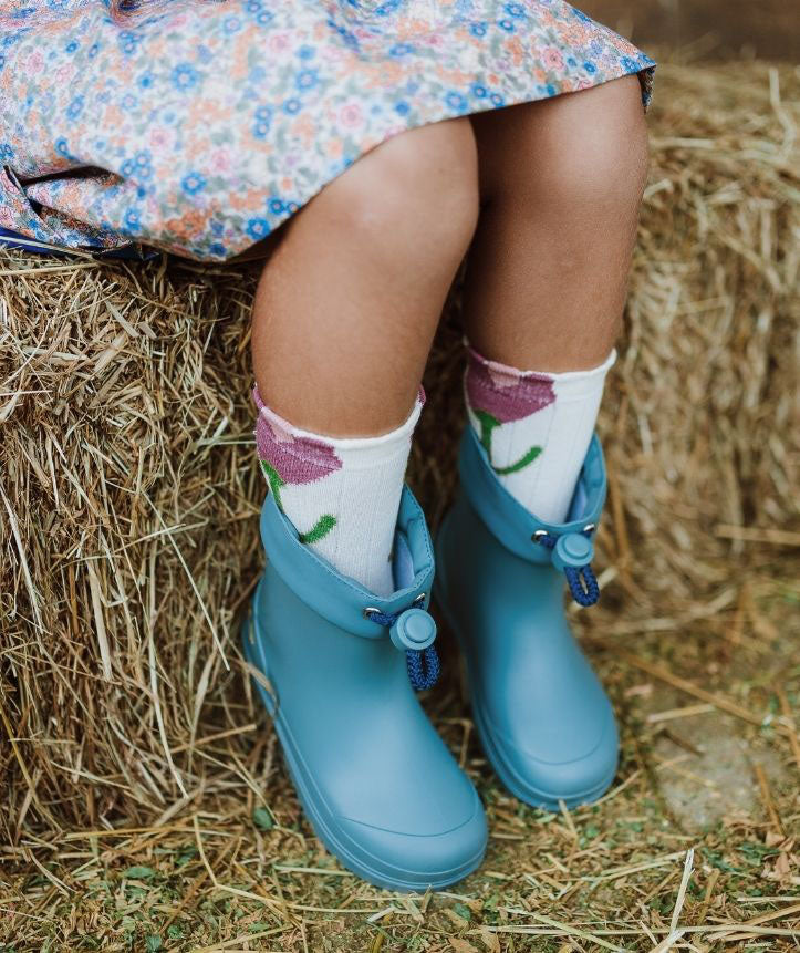 Pernas de uma criança a usar as galochas Igor MENDI Turquesa com meias coloridas, sentada numa pilha de feno.