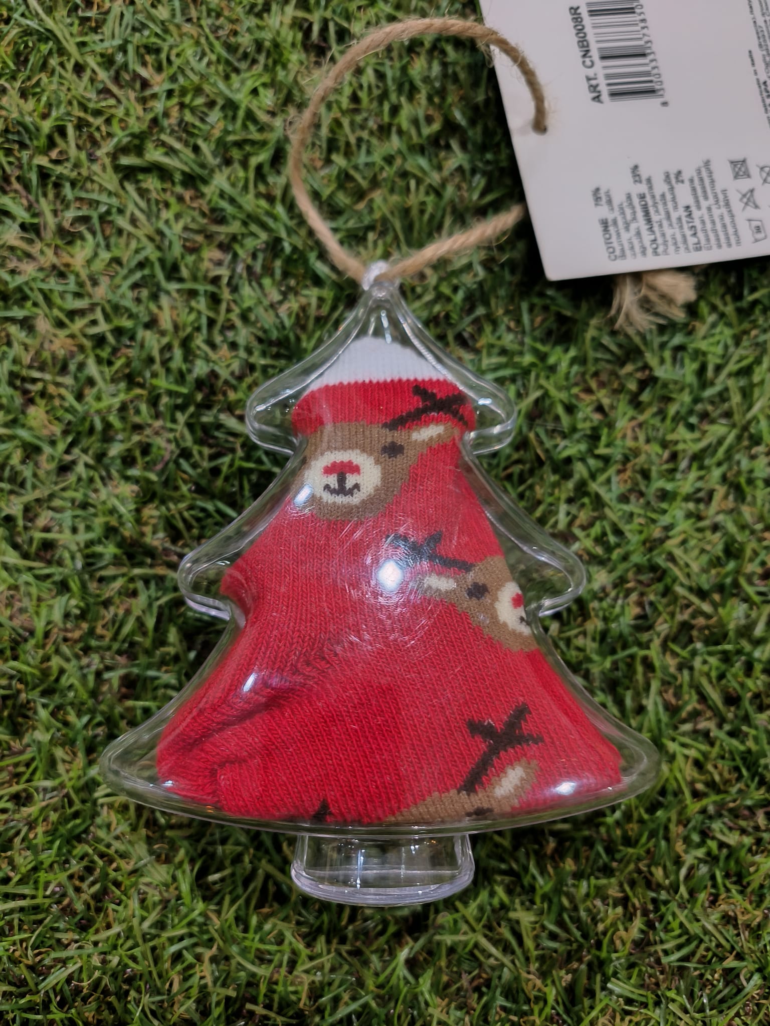 Meias de bebé Ciocca vermelhas com padrão de renas castanhas, dobradas dentro de uma caixinha transparente em forma de árvore de Natal, sobre um fundo de relva artificial.