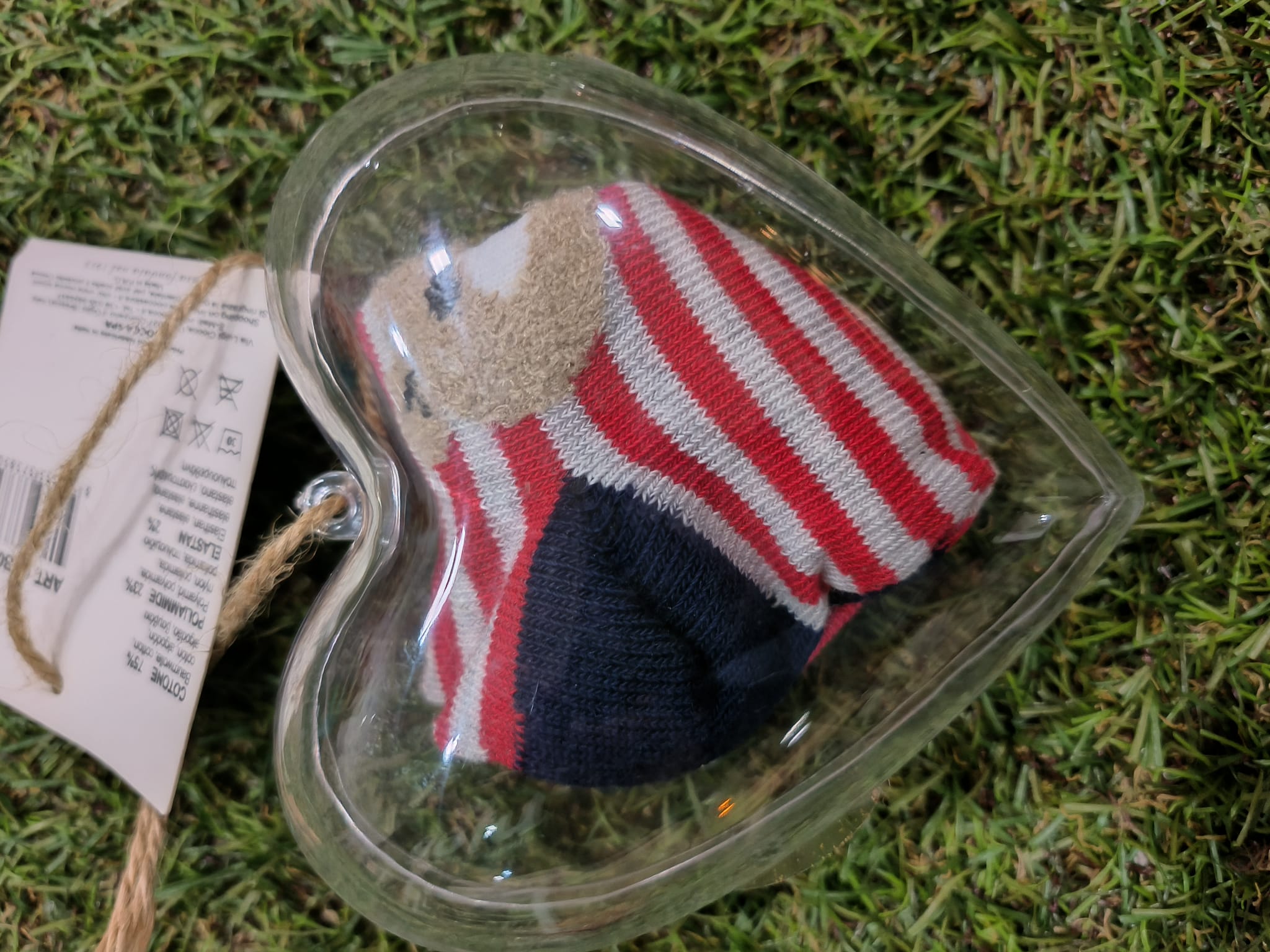 Meias de bebé Ciocca com riscas vermelhas, brancas e azuis, dobradas dentro de uma caixinha transparente em forma de coração, com uma etiqueta de composição visível, sobre um fundo de relva artificial.