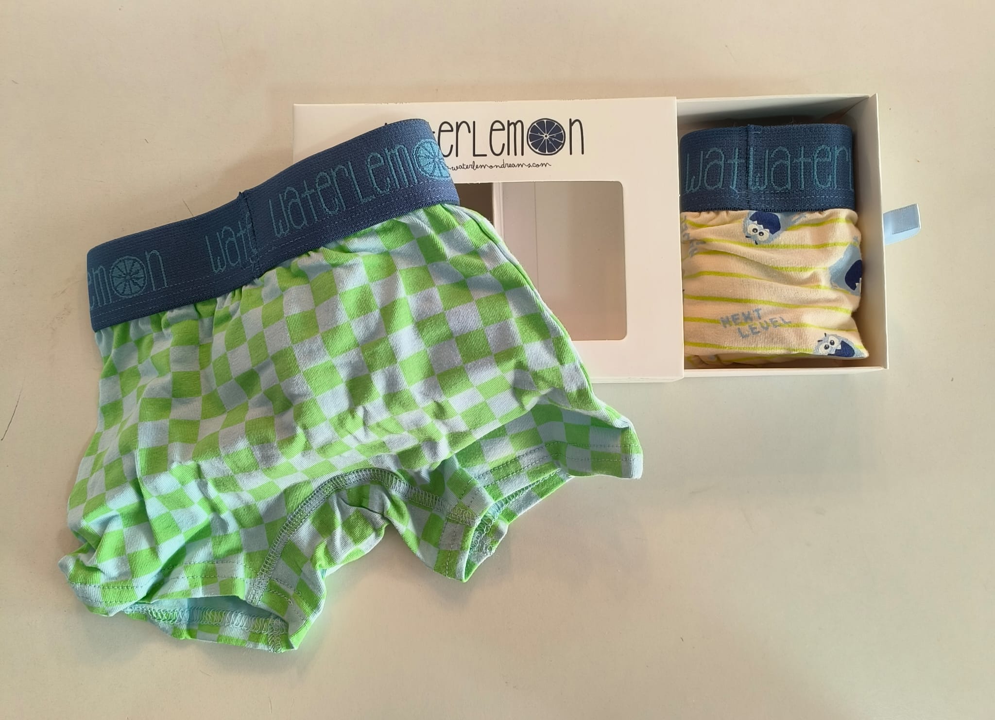 Boxer Waterlemon verde e azul xadrez e uma caixa aberta com outro boxer bege às riscas e com corujas.