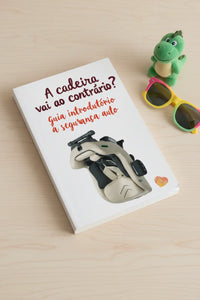 Capa do livro "A Cadeira vai ao contrário?" sobre uma mesa de madeira clara, com um brinquedo dinossauro e óculos de sol coloridos ao lado.