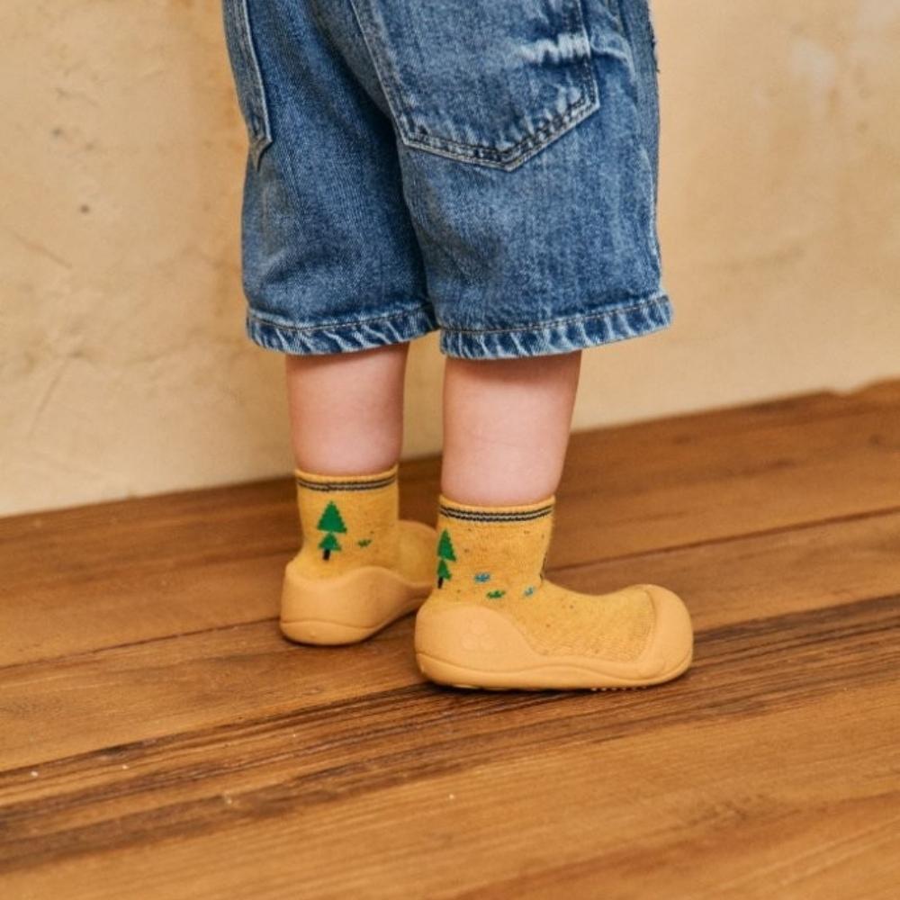 Pés de um bebé a usar os sapatos Attipas BICYCLE YELLOW, com padrão de árvores verdes, em pé sobre um chão de madeira.
