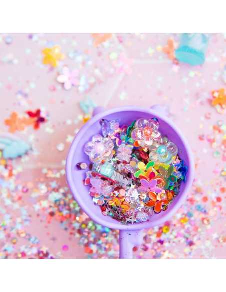 Detalhe de uma colher roxa cheia de brilhos coloridos e pequenas flores de plástico, ideal para atividades sensoriais.